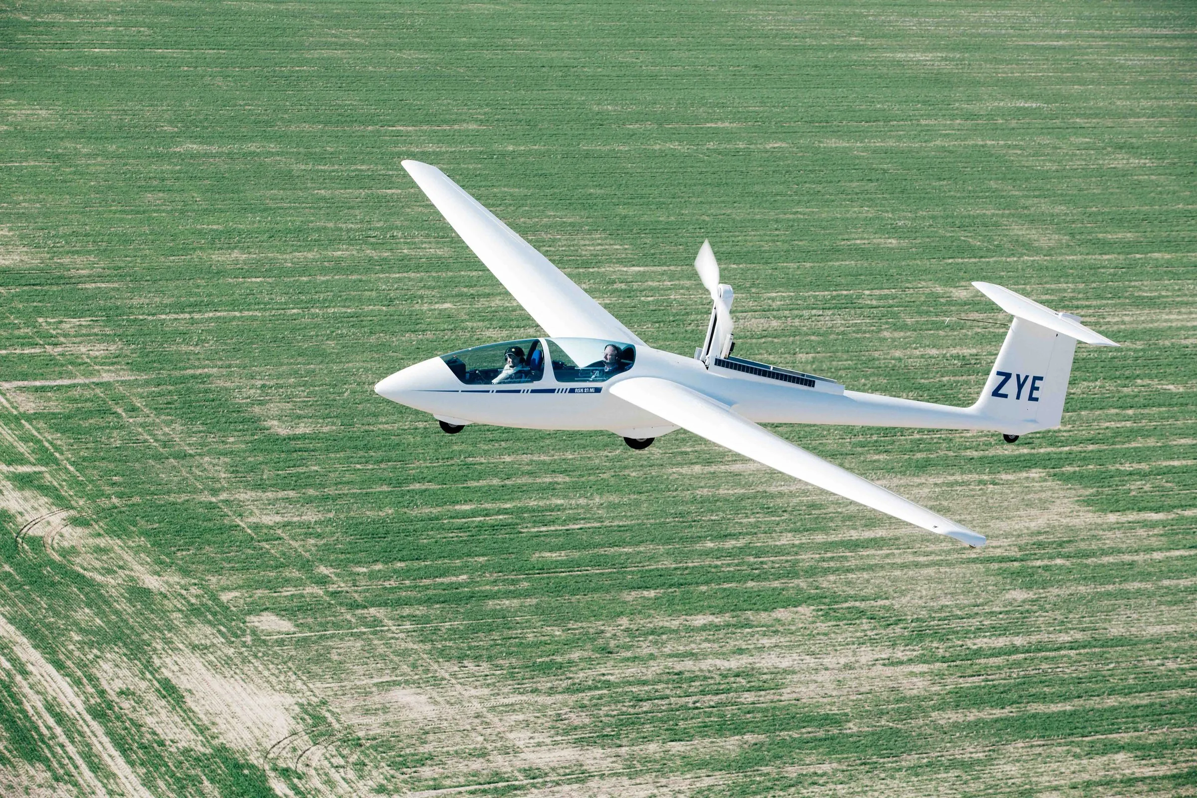 SoarAbility's motor glider VH-ZYE in flight over green wheatbelt crop, propeller deployed, pilot and instructor aboard