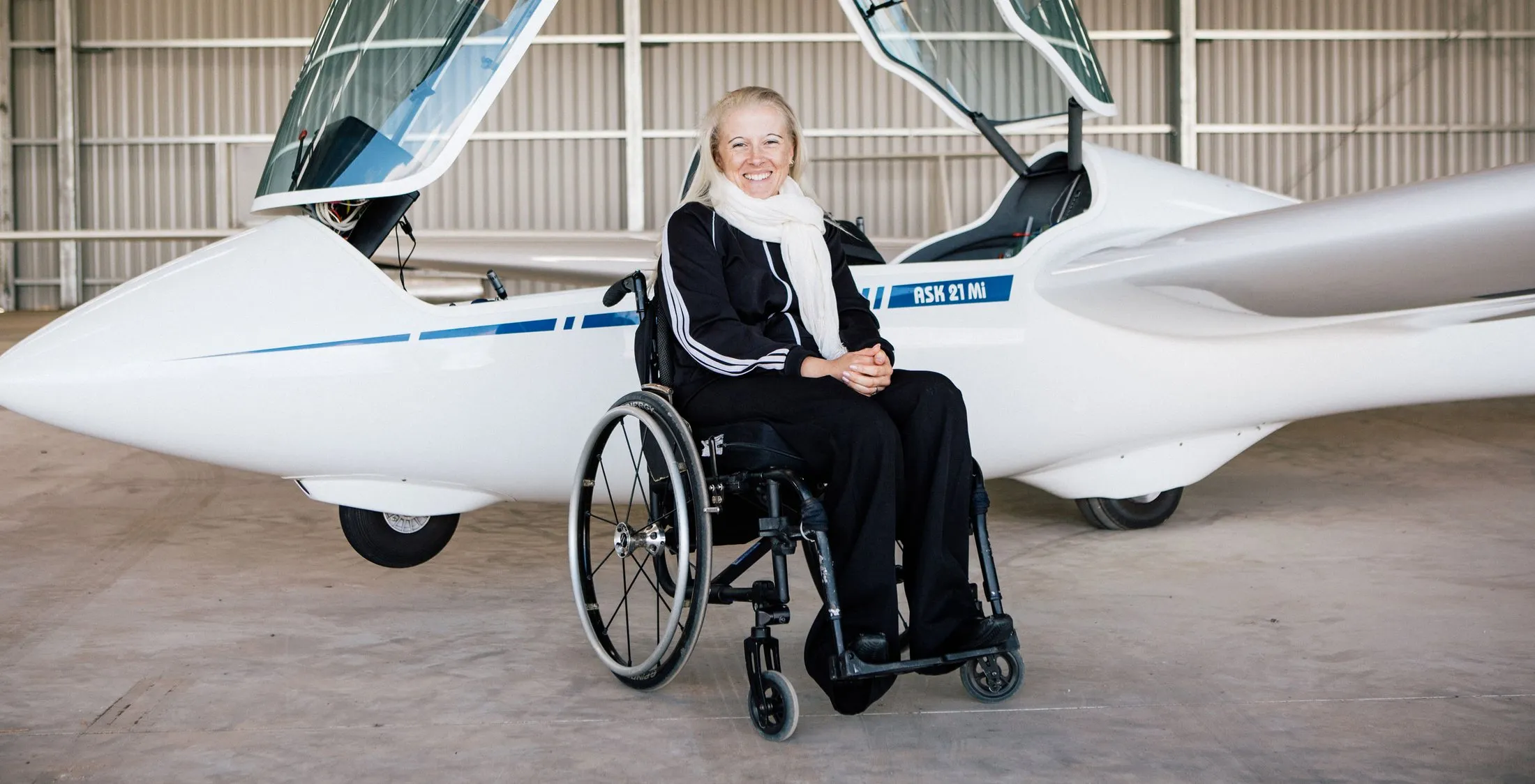 A woman in a wheelchair beside the open cockpit of SoarAbility's ASK21Mi in the hangar, smiling