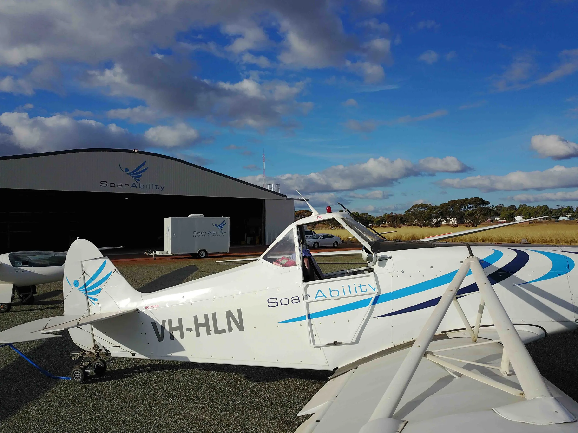 SoarAbility's white tow plane in its blue livery parked in front of the branded hangar under towering clouds